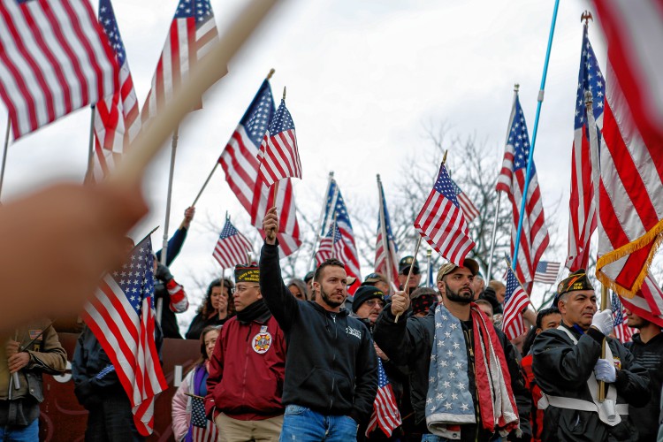 John Milbier Jr. of Springfield, son of a World World II veteran, cheers during a rally Sunday at Hampshire College to protest the school's decision not to fly the flag in the center of the Amherst campus. Below left, campus police officers Kate Godfrey, left, and Matt Brown form a barrier around a Hampshire student, center, who sat in front of the school's sign while protesters were trying to take a photo there. Below right, Tricia Pancione of Granby sings during the protest. GAZETTE STAFF/SARAH CROSBY